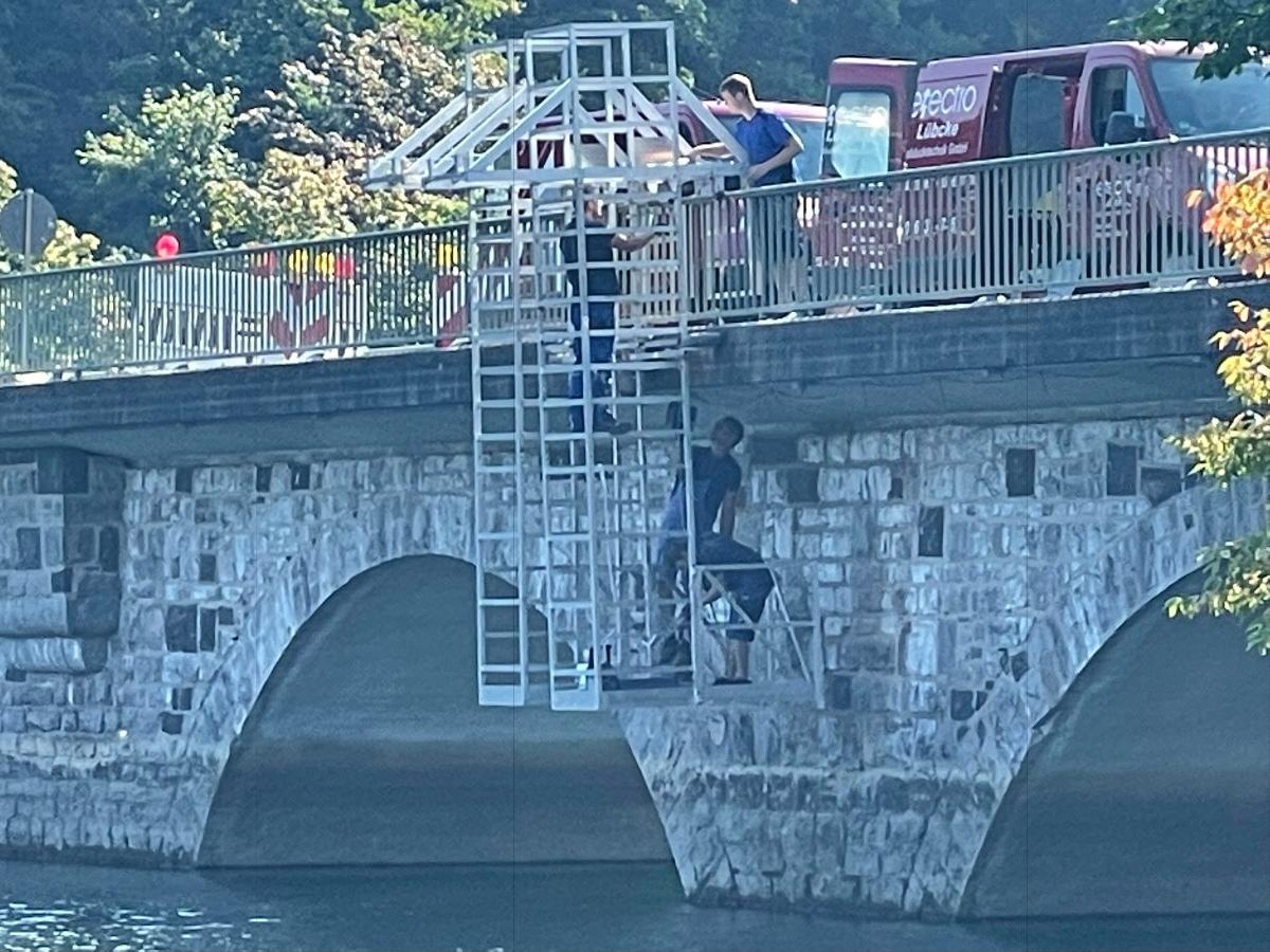 Monteure arbeiten von einem Hängegerüst aus am Brückenpfeiler, auf der Brücke steht das rote Firmenfahrzeug.