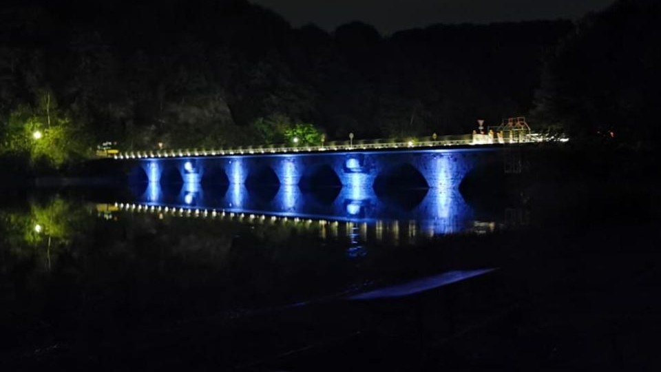 Die Steinbogenbrücke bei Nacht, blau angestrahlt, das Licht spiegelt sich im Wasser.