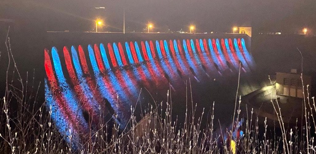 Die Sperrmauer in voller Breite, jeder Mauerbogen ist einzeln rot oder blau angestrahlt.