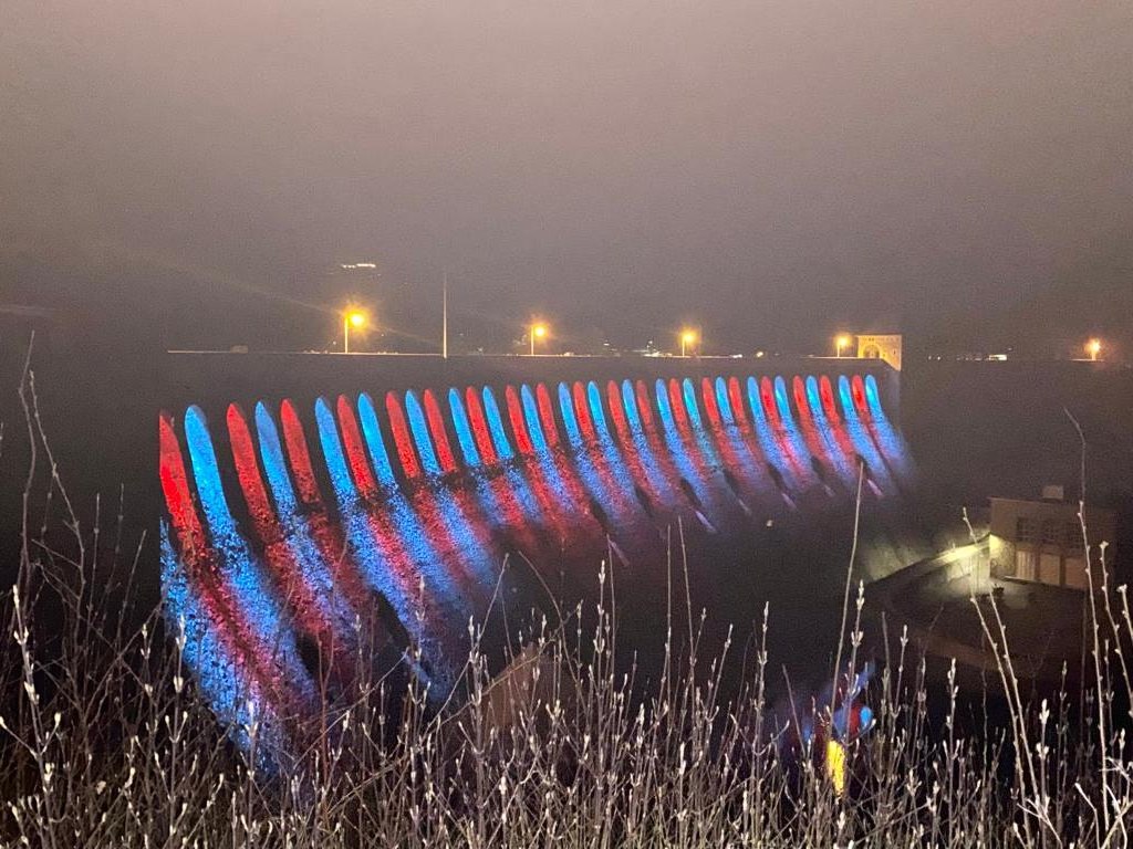 Die Edersee-Sperrmauer bei Nacht und Nebel, die Mauerbögen leuchten abwechselnd rot und blau.